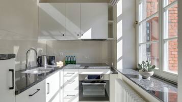 Une cuisine moderne avec des armoires blanches, un four encastré, un évier en acier inoxydable et une fenêtre donnant sur un bâtiment en briques.