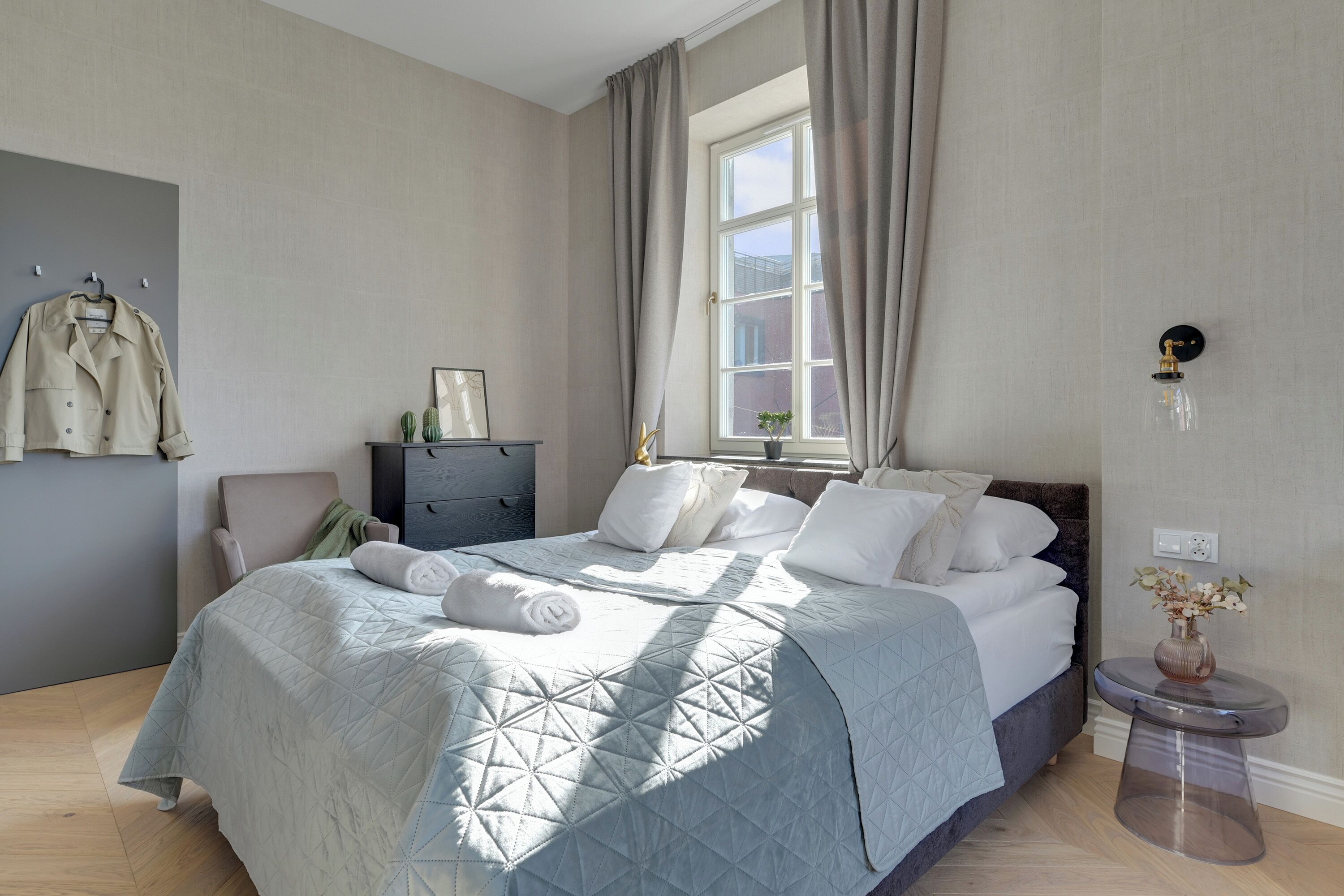 Un dormitorio con cama, cómoda, silla, ventana con cortinas e iluminación en la pared.
