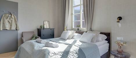 Un dormitorio con cama, cómoda, silla, ventana con cortinas e iluminación en la pared.
