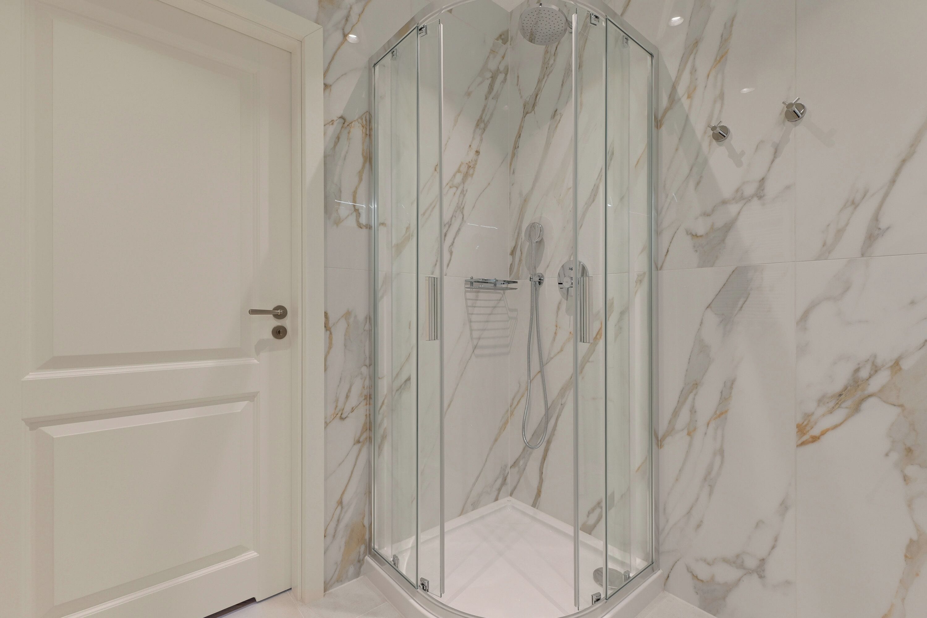 A glass-enclosed shower with a white door and marble-patterned walls.