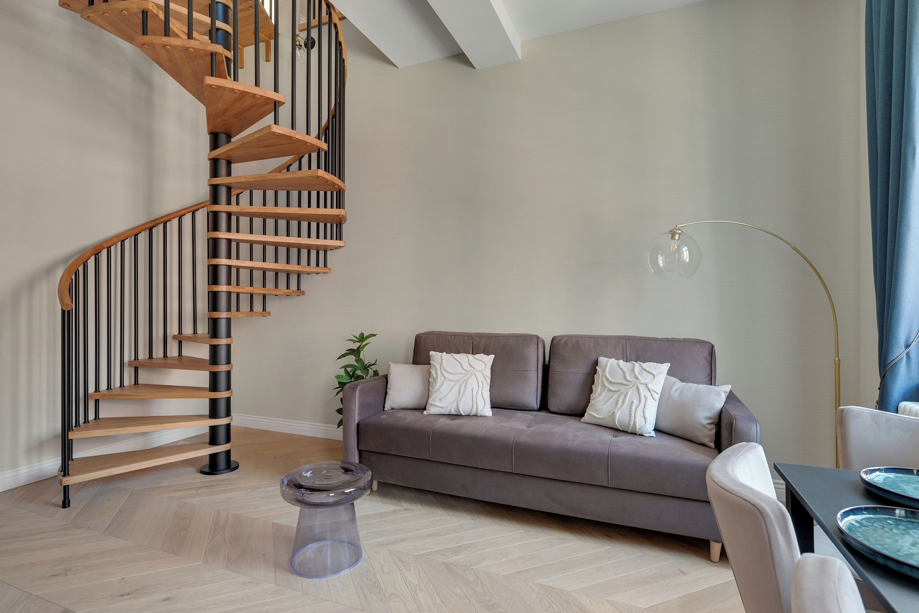 A living room with a wooden spiral staircase, a grey sofa, a floor lamp, and a dining table.