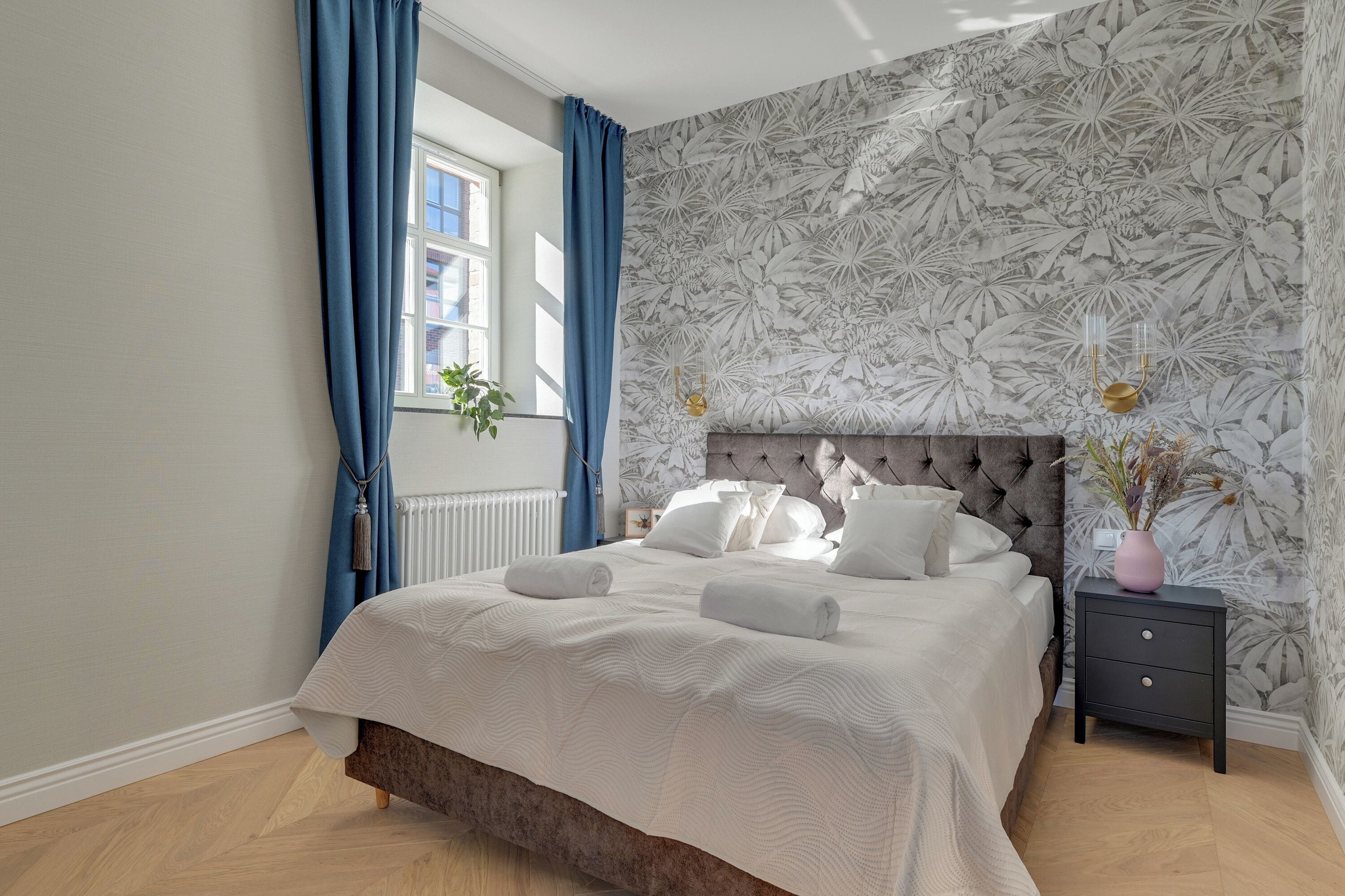 Un dormitorio con cama, mesita de noche, ventana con cortinas y papel tapiz con motivos florales.