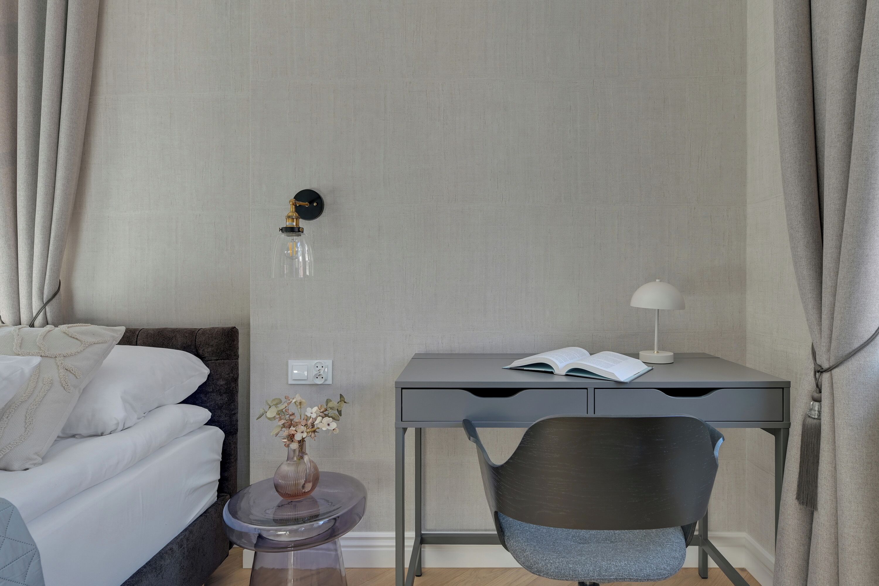 A hotel room with a bed, a desk with a lamp, a chair, and a small table with a vase.