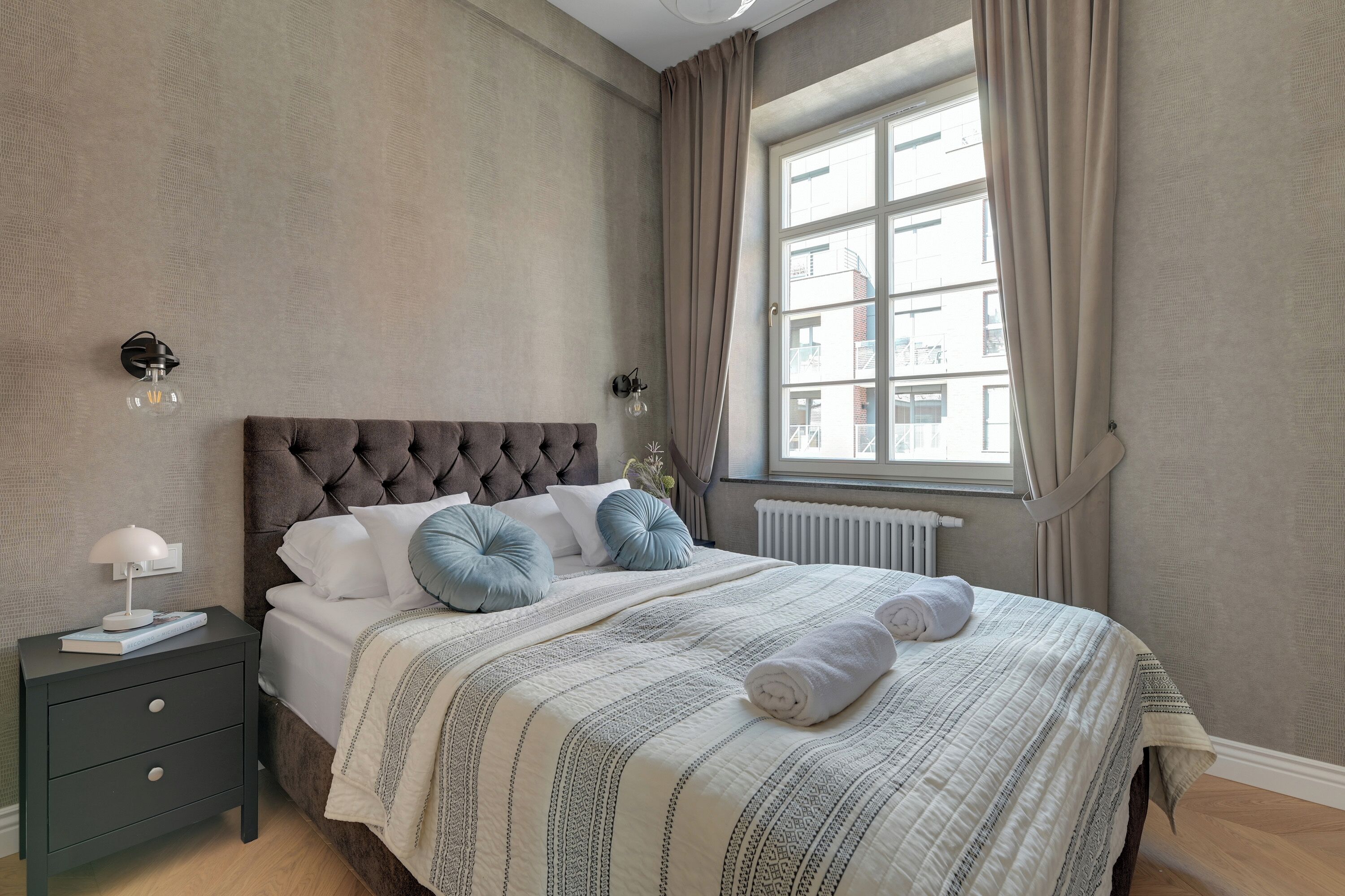 Una camera da letto con testiera trapuntata, un letto con biancheria a righe, un comodino con una lampada e una finestra con tende.