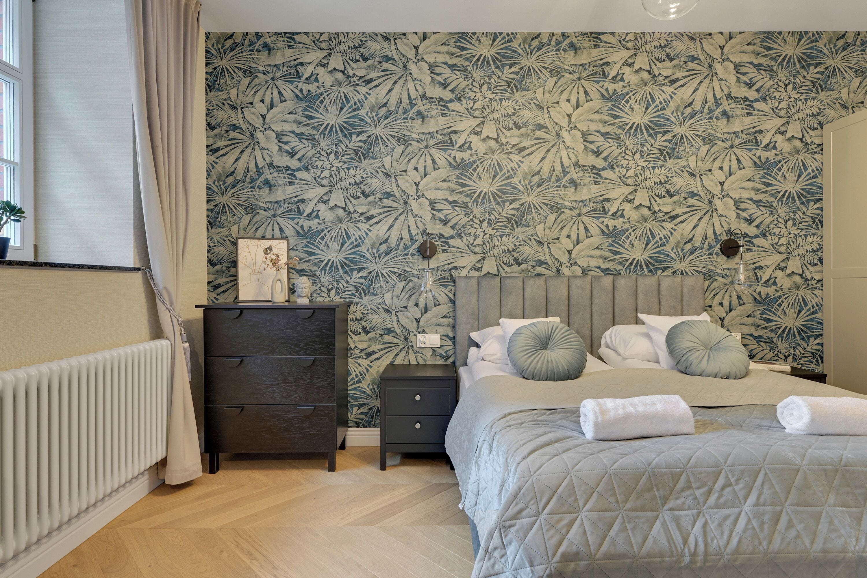 Et soveværelse med blomstret tapet, en seng med puder, et natbord, en kommode og en radiator.