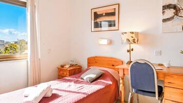 A bedroom with a bed, a desk, a chair, a lamp, and a window with a view of buildings.