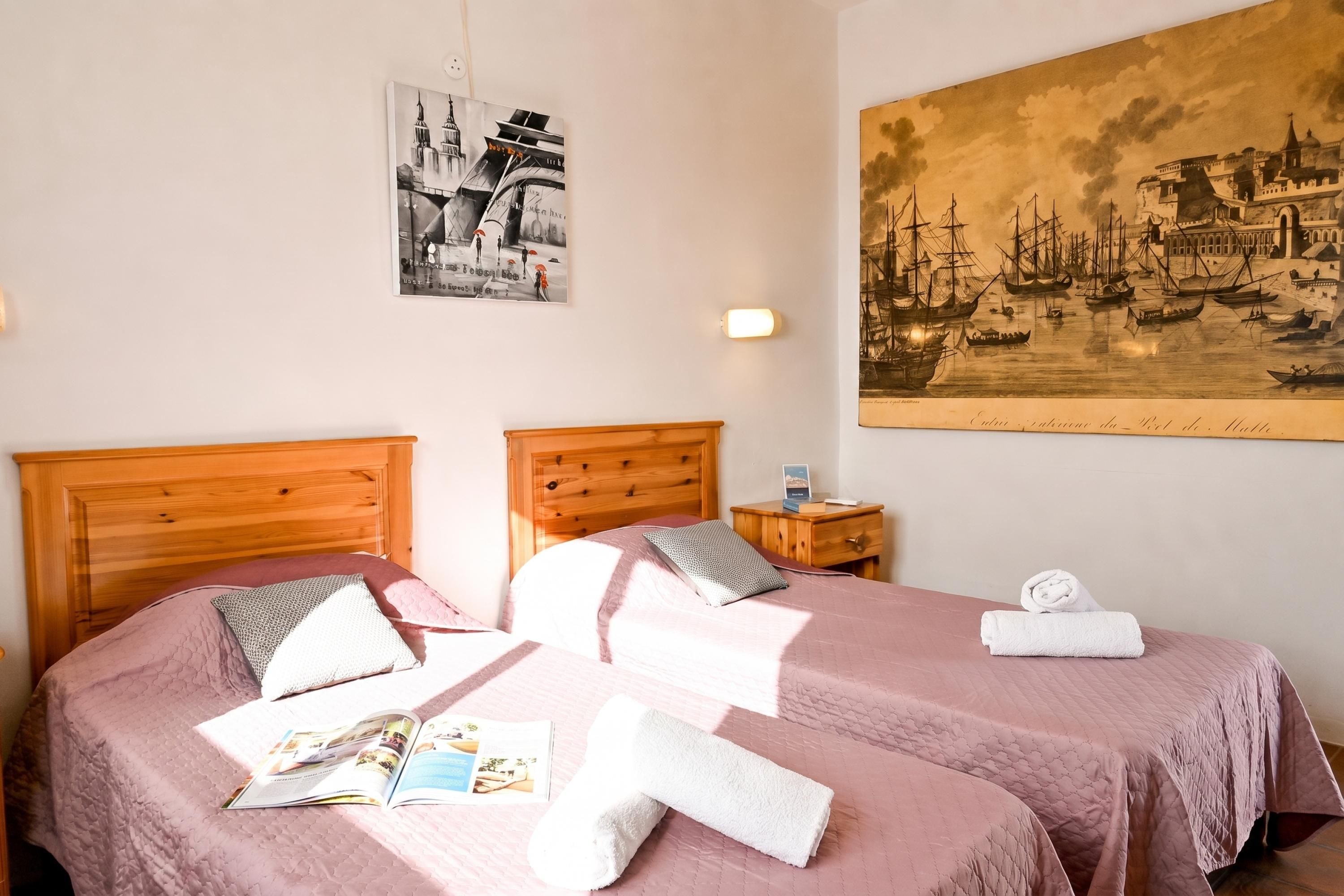 Una habitación de hotel con dos camas, una mesita de noche, una lámpara fijada a la pared y un cuadro histórico grande.