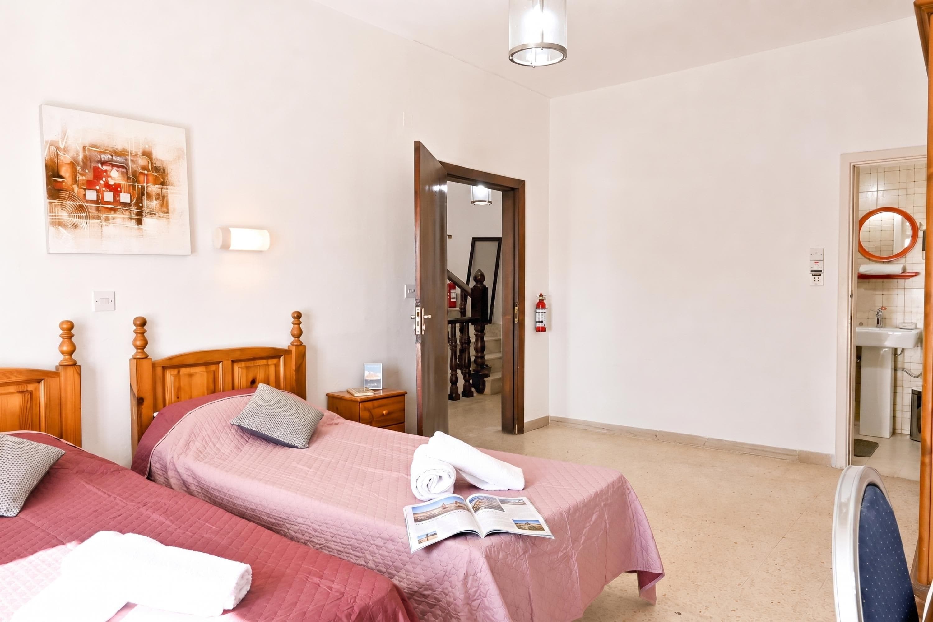 A hotel room with two beds, a bedside table, a mirror, a painting, and a door leading to a bathroom.