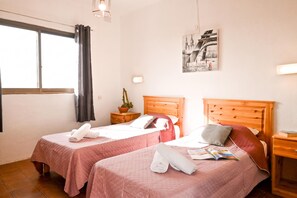 Habitación de hotel con dos camas, una ventana con cortinas, una lámpara de pared, una planta en maceta y un cuadro enmarcado.