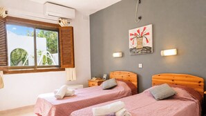 Habitación de hotel con dos camas, ventana con persianas, equipo de aire acondicionado, una obra de arte en la pared y una mesita de noche con lámpara.