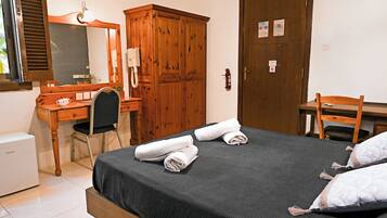 Un dormitorio con cama, armario de madera, escritorio con silla, espejo y un refrigerador pequeño.