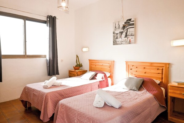 Une chambre d’hôtel avec deux lits, une fenêtre avec des rideaux, une table de chevet avec une plante, une lampe fixée au mur et un tableau encadr é.