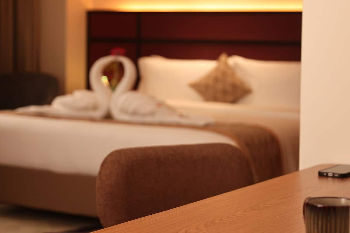 A hotel room with a wooden table, a bed with white linens, a headboard, and a hanging lamp.