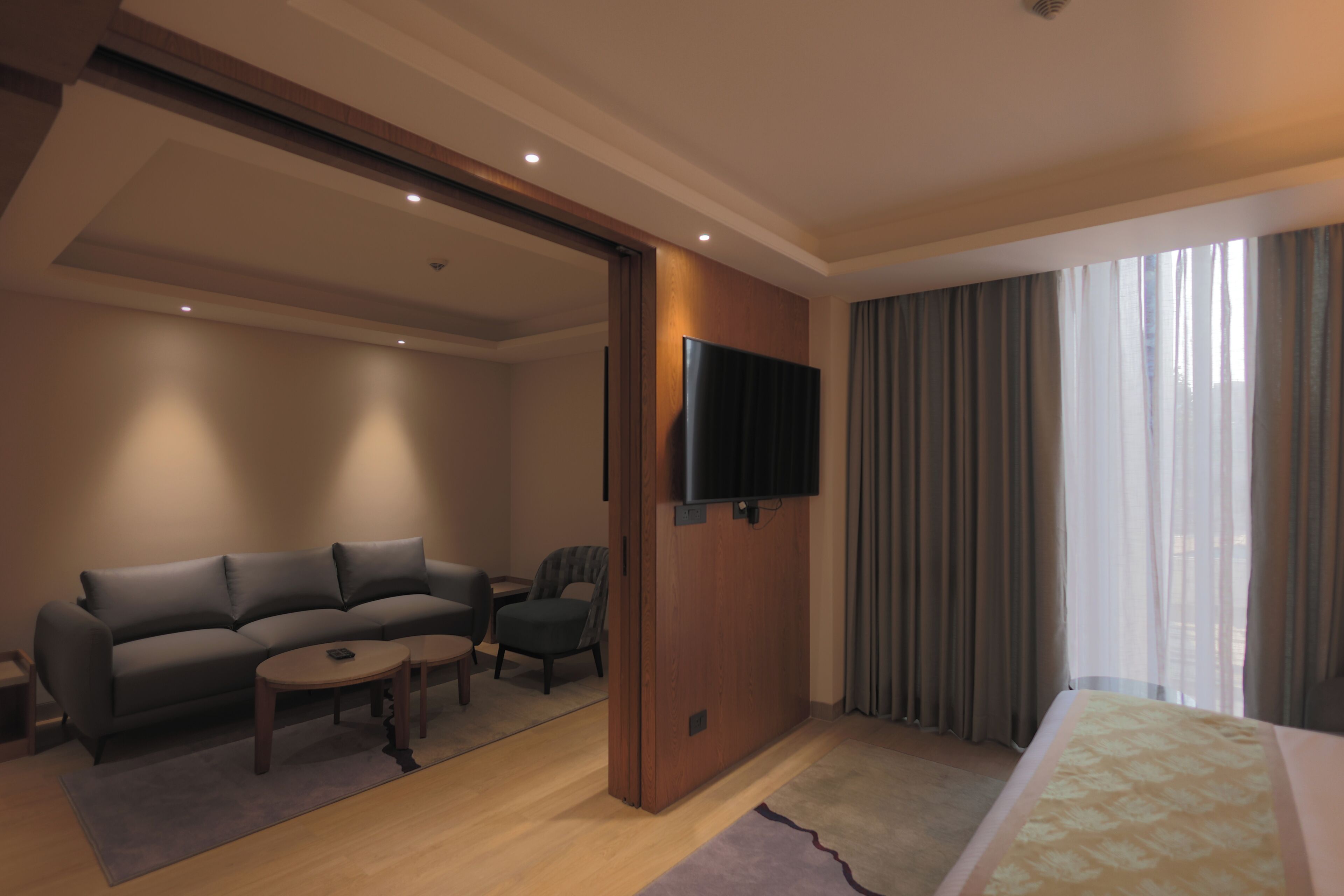 Quarto de hotel com sofá, poltrona, mesinha, TV fixada em um painel de madeira e cama com colcha estampada.