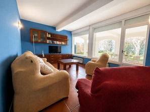 A living room with a sofa, armchair, TV, bookshelf, and large windows.