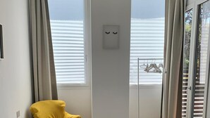 Une chambre d’hôtel avec un lit, un fauteuil jaune, une fenêtre avec des stores et une vue sur la mer.