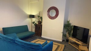 Un salon avec un canapé de couleur turquoise, un téléviseur posé sur un meuble en bois, un miroir au mur et une plante.