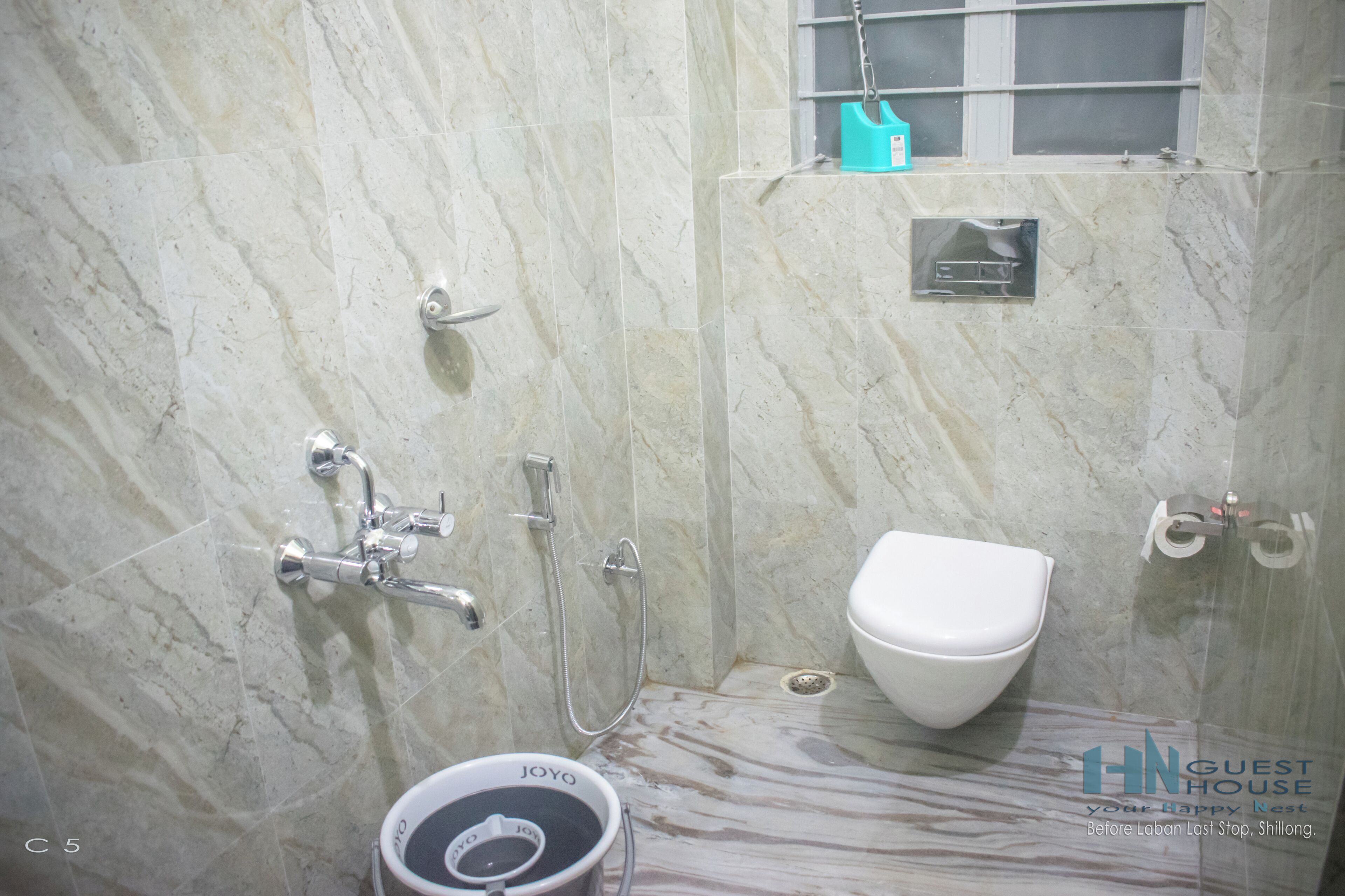 A bathroom with a marble floor, a white toilet, a shower with a handheld showerhead, and a small window above the toilet.