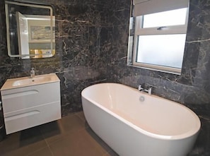 A bathroom with a marble wall, a white bathtub, a sink with a mirror, and a window.