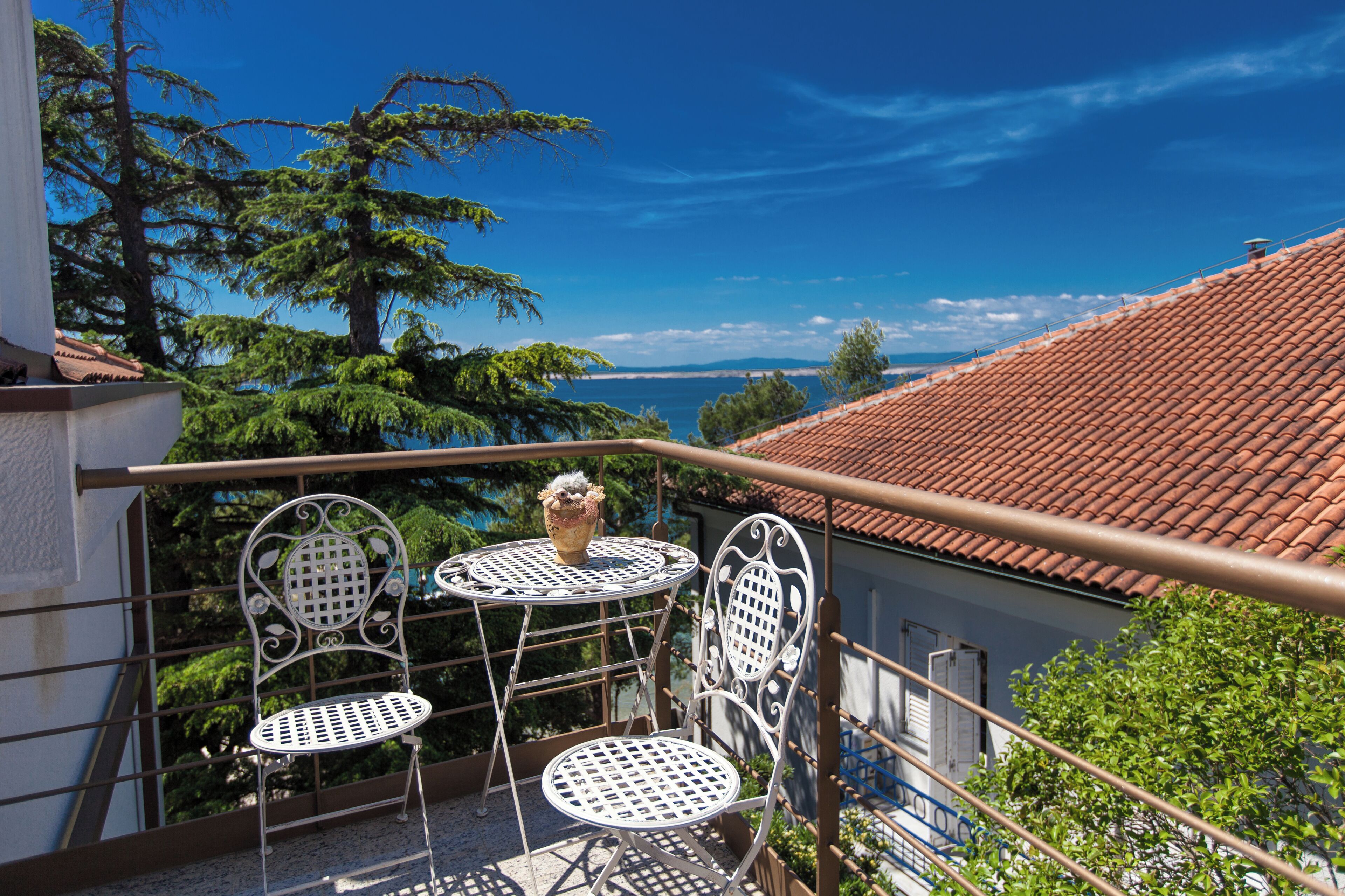 Un balcon avec deux chaises en métal et une table, offrant une vue sur la côte, avec des arbres et un toit recouvert de tuiles.