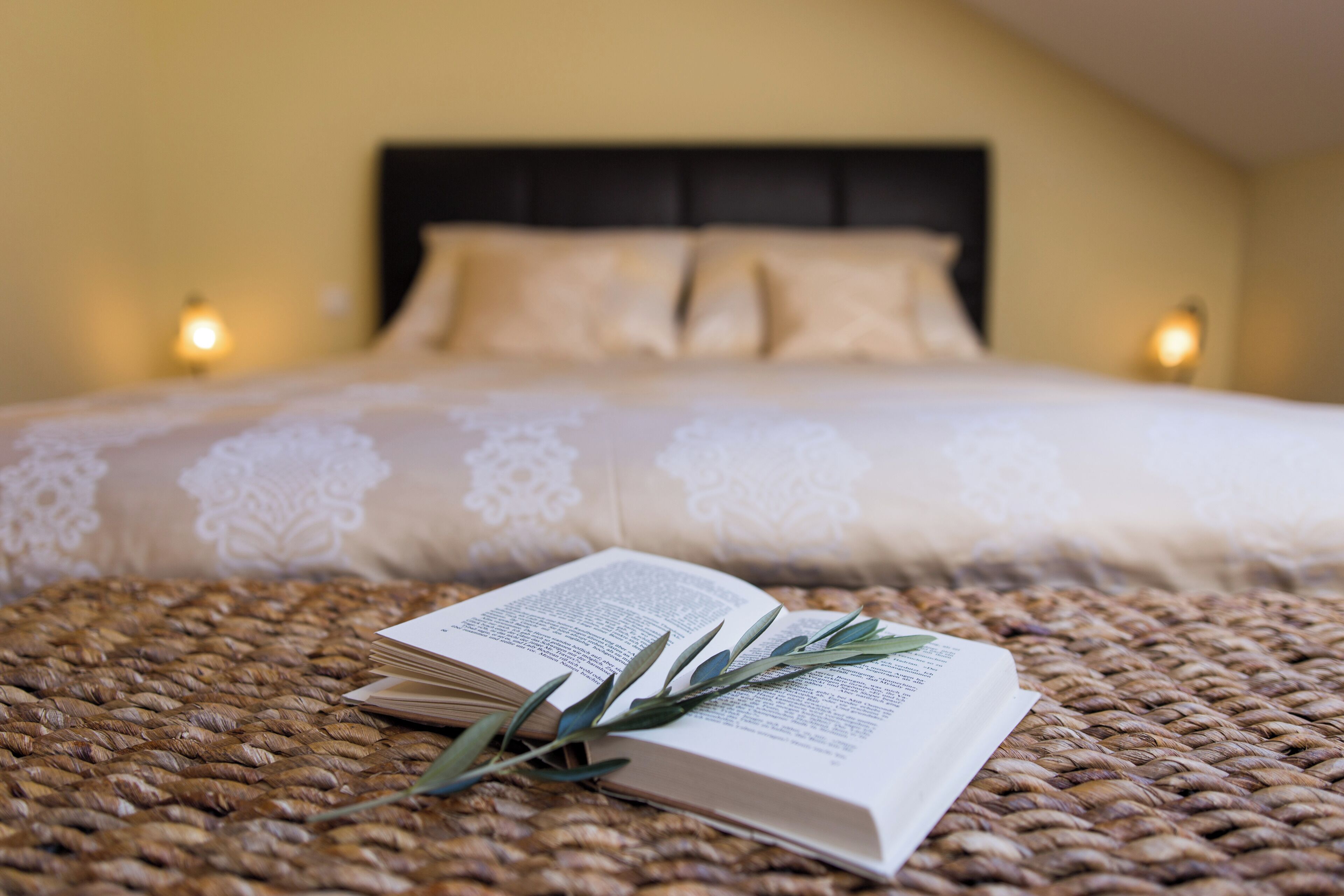 A bed with a white bedspread, a book, and a branch on a woven surface.