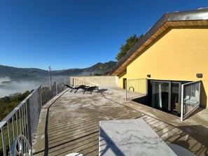 Eine Holzterrasse mit einem gelben Gebäude, einer Glastür und Blick auf Hügel.