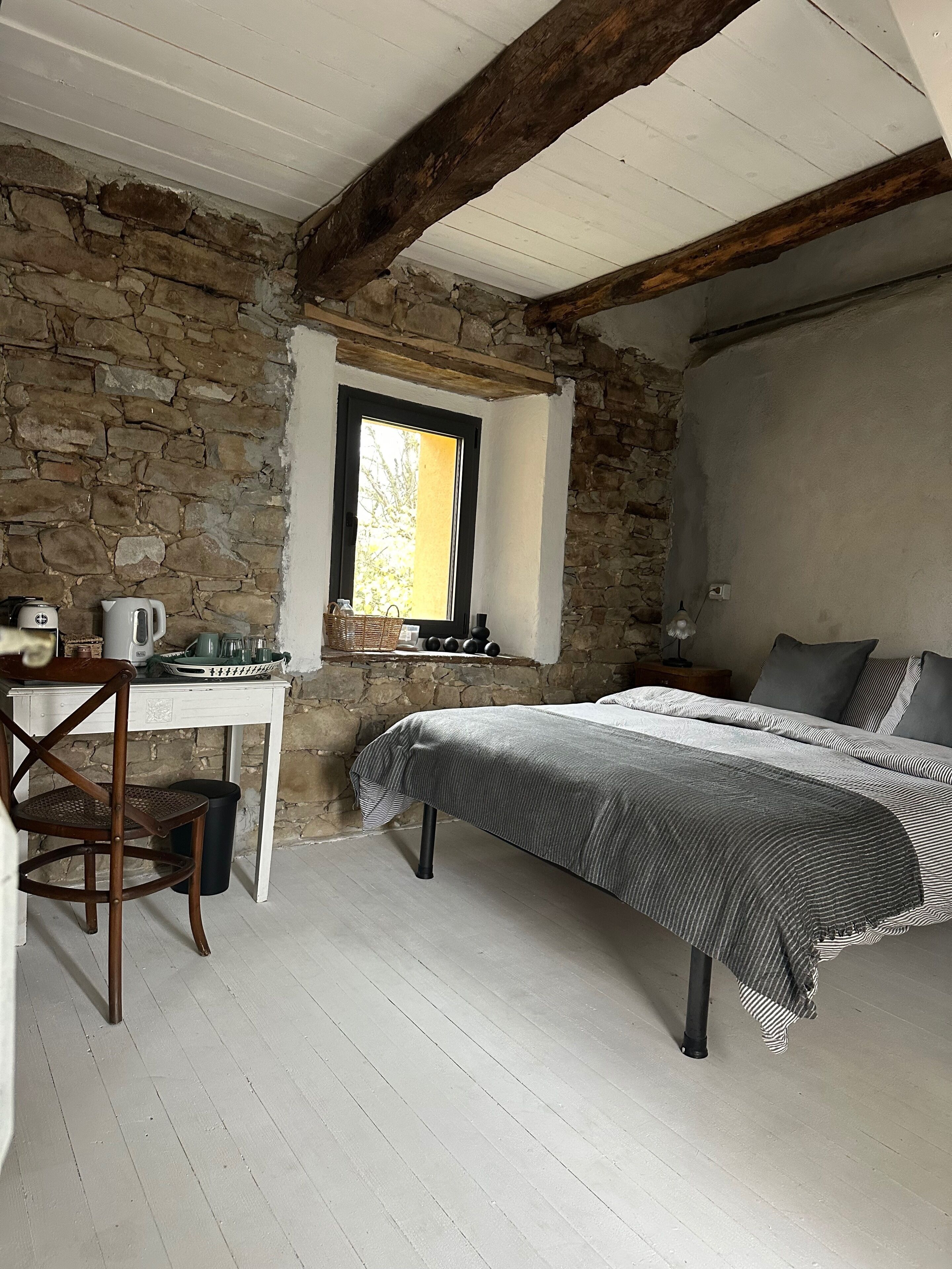 A bedroom with a stone wall, a wooden beam ceiling, a bed with a striped bedspread, a small table with a kettle, and a chair.