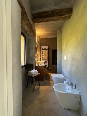 Ein schmaler Flur führt zu einem Badezimmer mit einer Badewanne, einer Toilette und einem Waschbecken. Am linken Rand befindet sich ein Stuhl mit Handtüchern und ein Fenster.