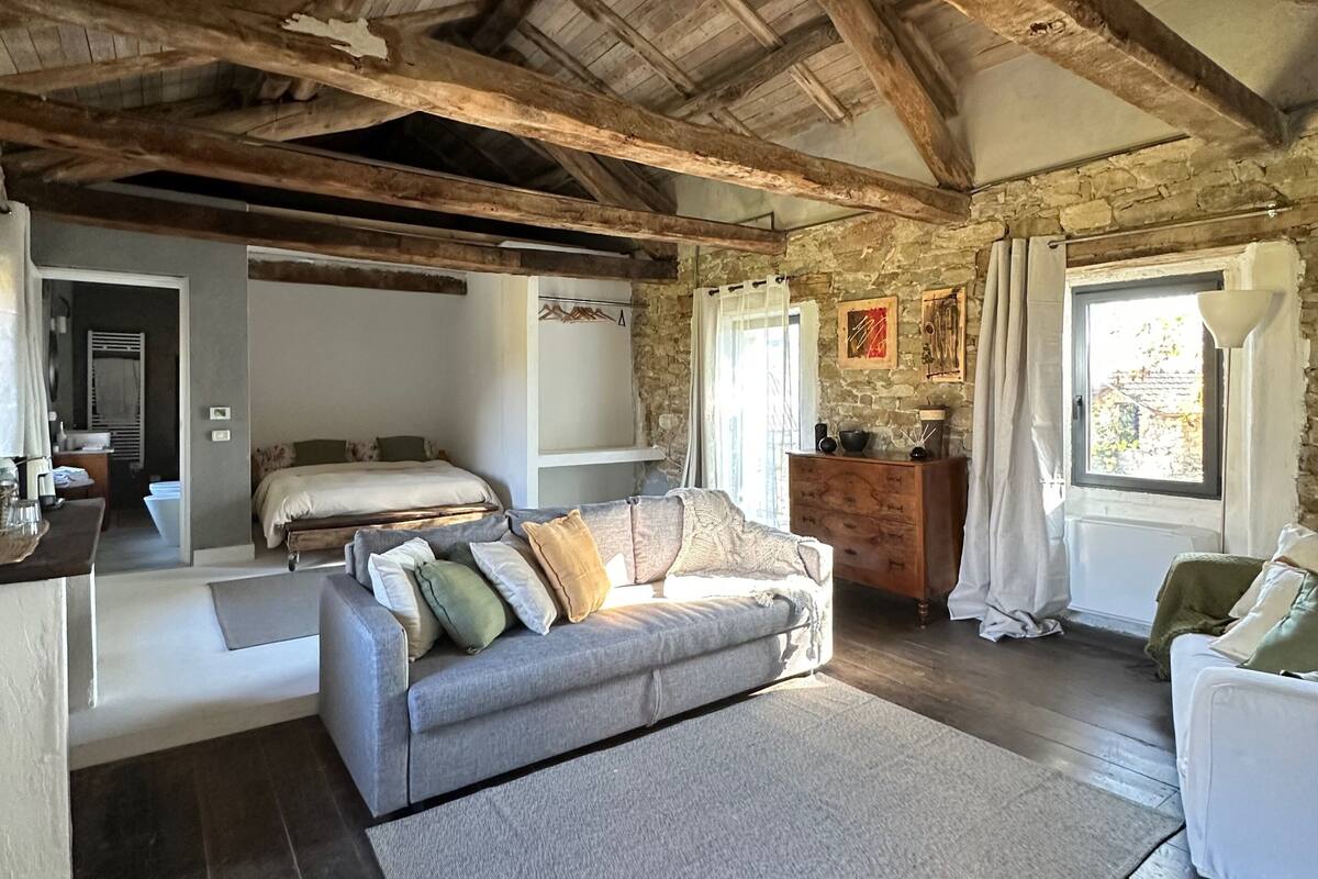 Un interno rustico con travi a vista in legno, un divano grigio con cuscini, una cassettiera bianca e una parete in pietra.