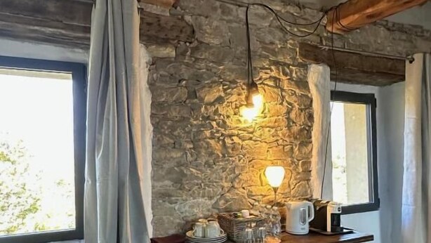 Una camera rustica con pareti in pietra, soffitto in legno, una finestra con tende e uno scaffale con oggetti da cucina.