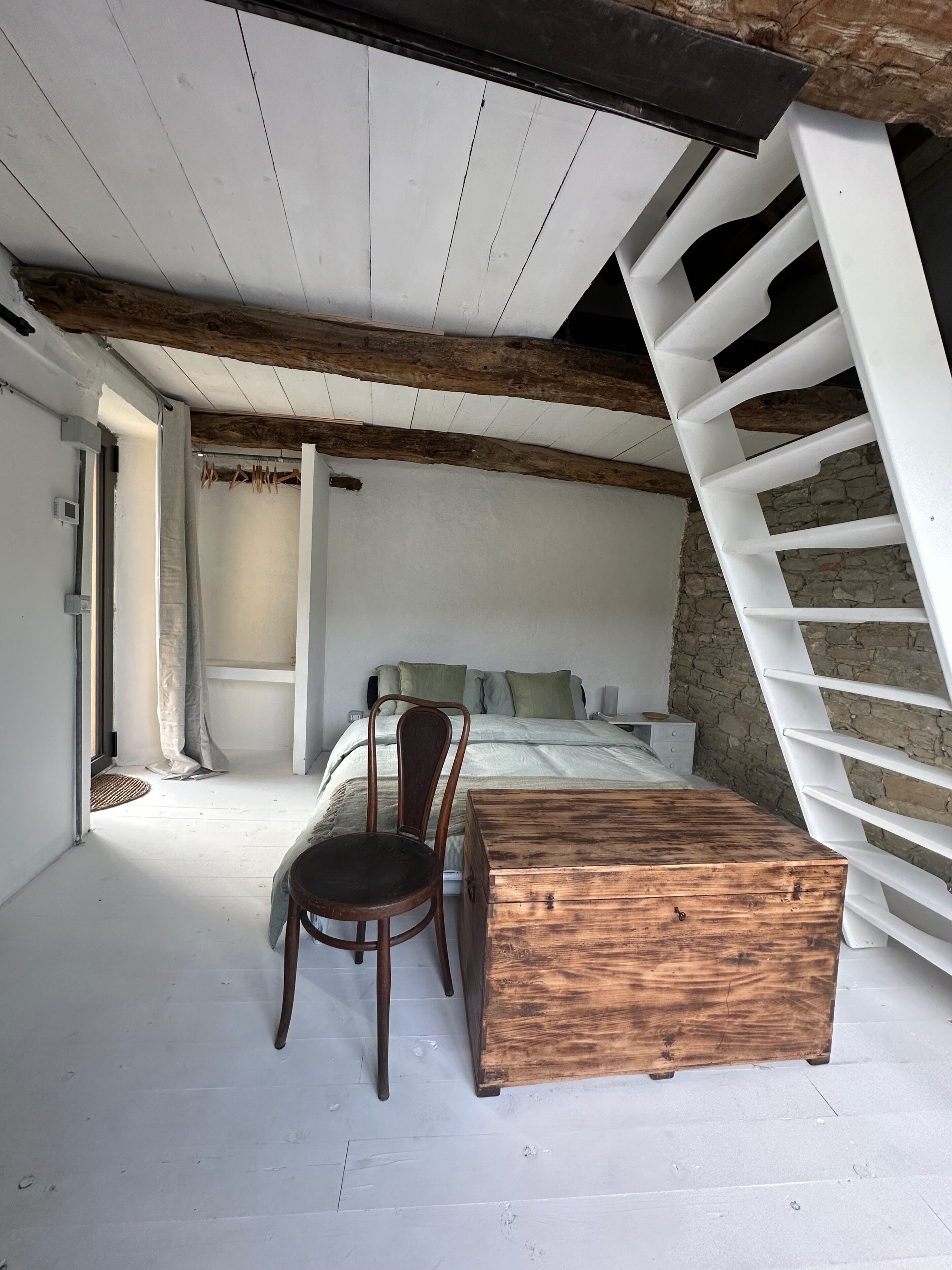A bedroom with a bed, a wooden chest, a chair, and a white staircase.