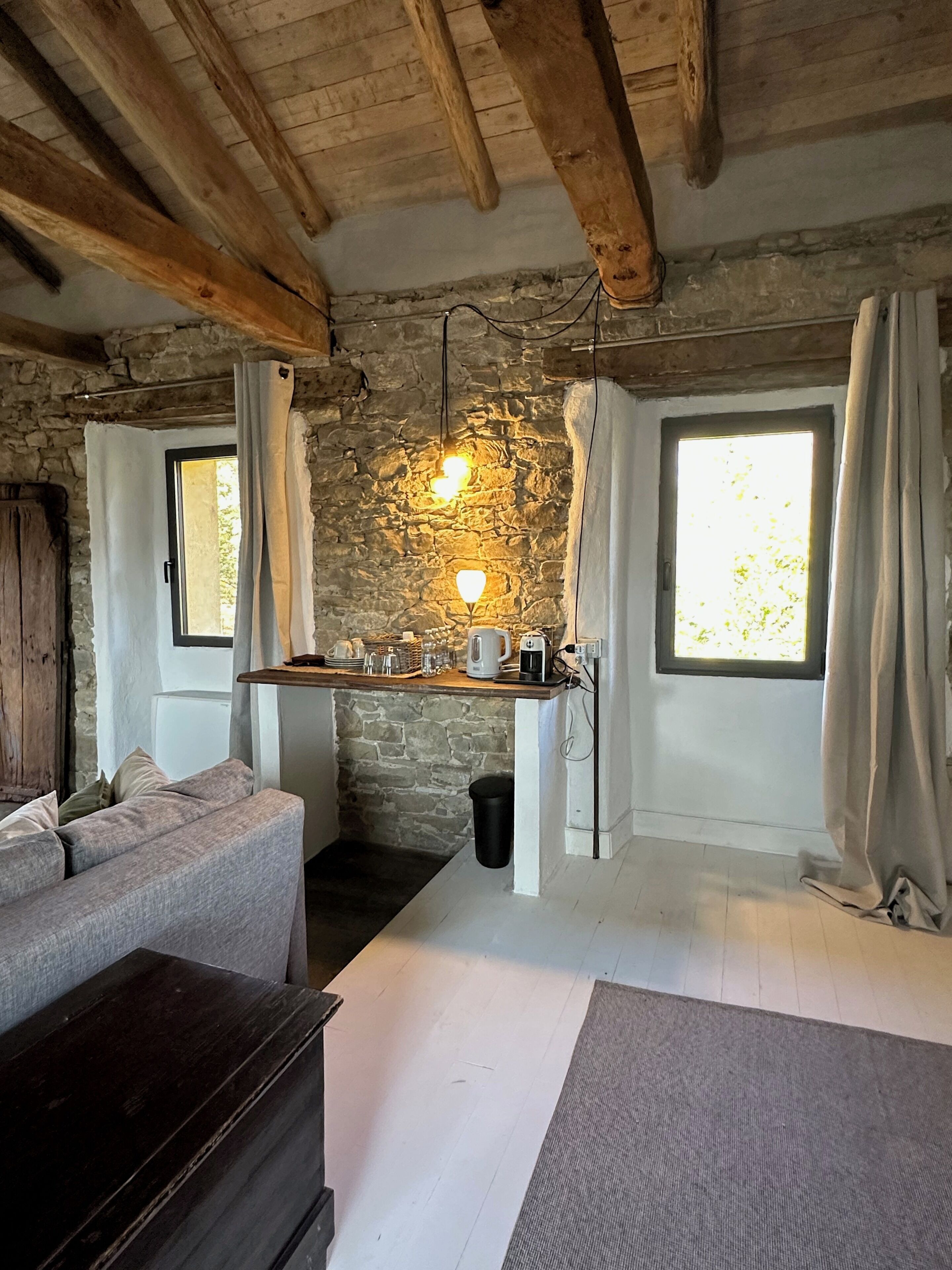 Ein rustikaler Raum mit freiliegenden Steinwänden, Holzbalken, einer Couch, einem kleinen Tisch mit Gläsern und einem Wasserkocher sowie einem Fenster mit Vorhängen.
