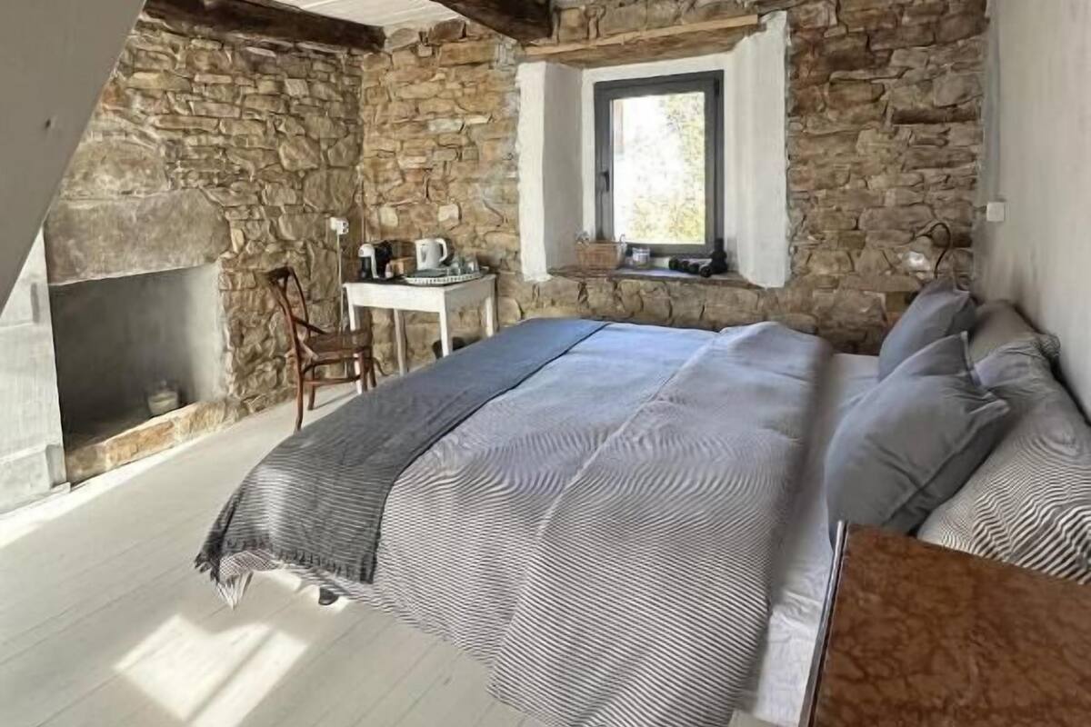 Una camera da letto con parete in pietra, soffitto in legno, un letto con copriletto a righe, un piccolo tavolo con lampada e una finestra.
