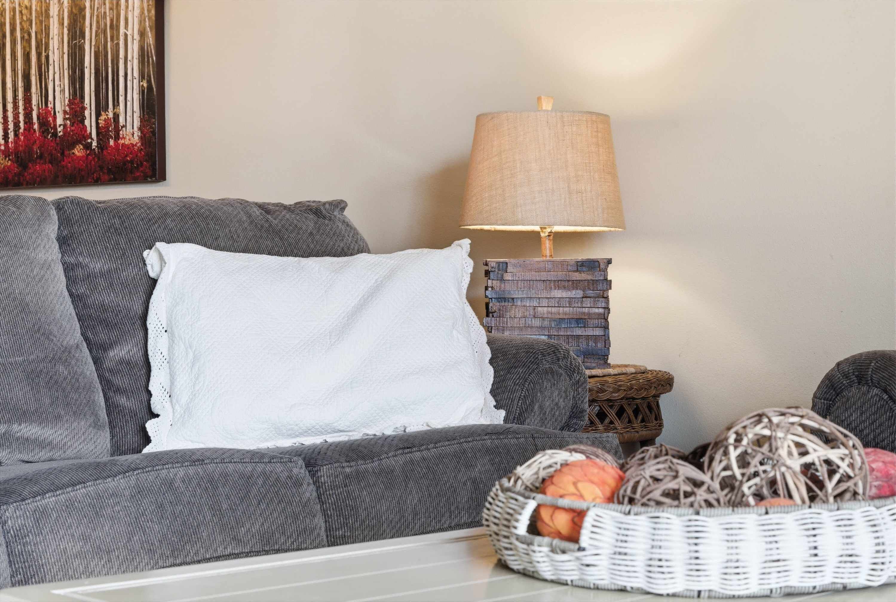 A grey sofa with a white pillow, a lamp, a basket with fruits, and a painting on the wall.