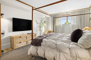 A bedroom with a four-poster bed, a television on the wall, a bedside table, a lamp, and a window with curtains.