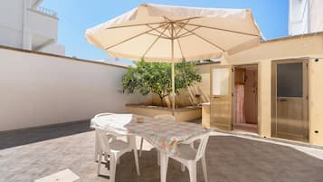 En udendørs terrasse med et bord, stole, en beige parasol og en potteplante.