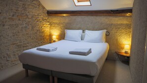 A bedroom with a stone wall, a wooden beam ceiling, a skylight, a bed with white linens, two bedside lamps, and a small bedside table. - L'Ancien Relais (Genouilly)
