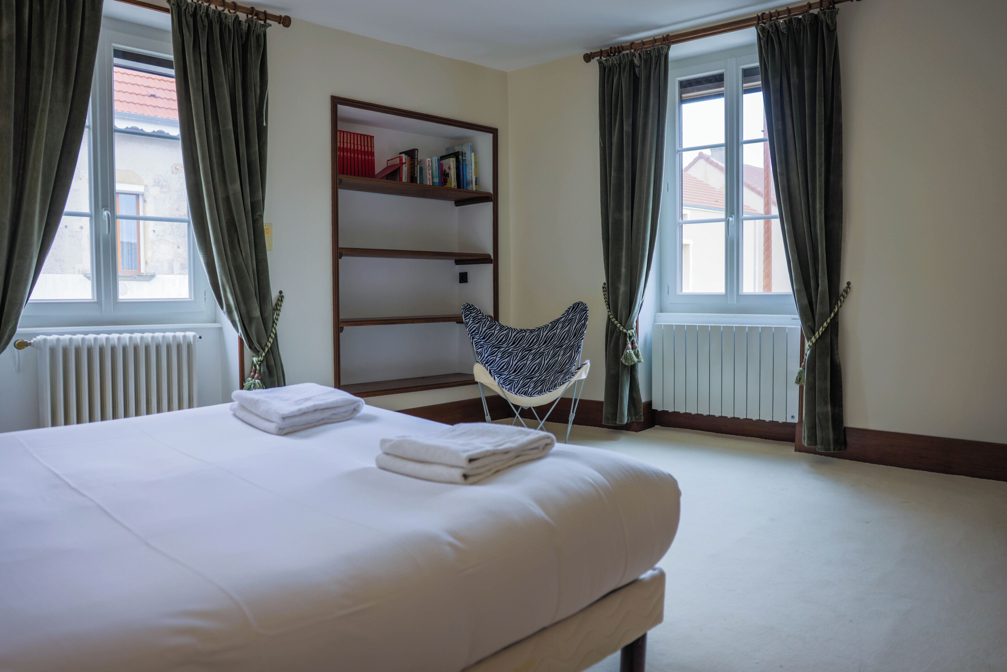A hotel room with a bed, a chair, a bookshelf, and a window.