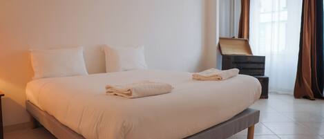 A large bed with a grey frame, white bedding, and two pillows. There is a small wooden nightstand with a lamp and a box on it.
