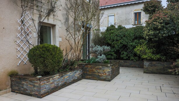 A courtyard with tiled flooring, stone planters, and a building with a window and door. - L'Ancien Relais (Genouilly)