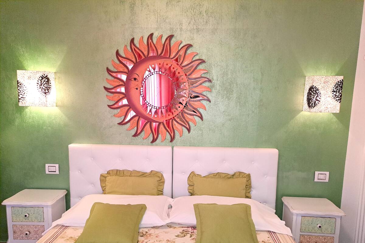 A room with a bed, two nightstands, a wall-mounted decorative mirror, and two wall sconces.