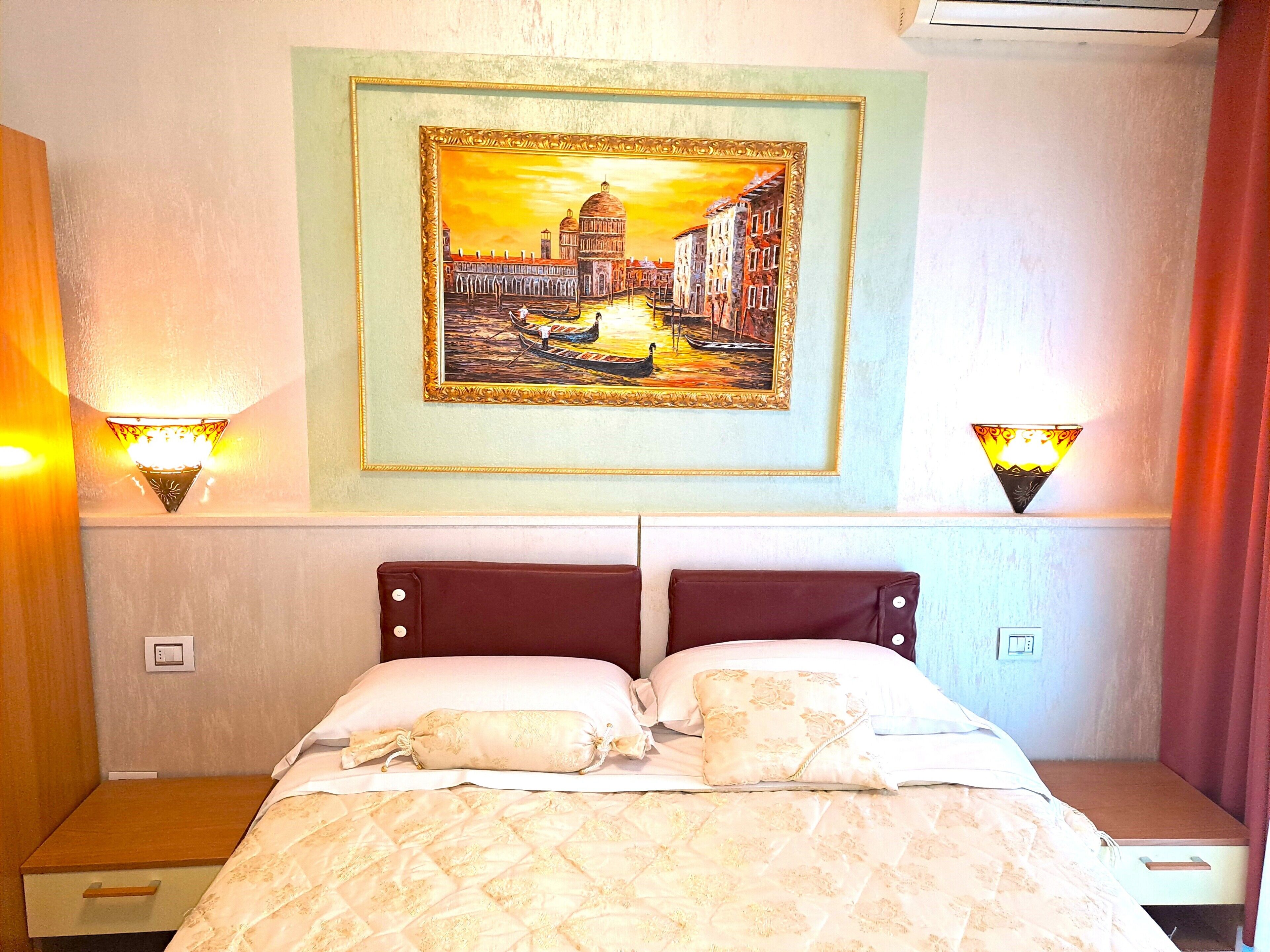 A hotel room with a bed, two wall-mounted lamps, a framed painting of a cityscape, and a nightstand with a lamp.