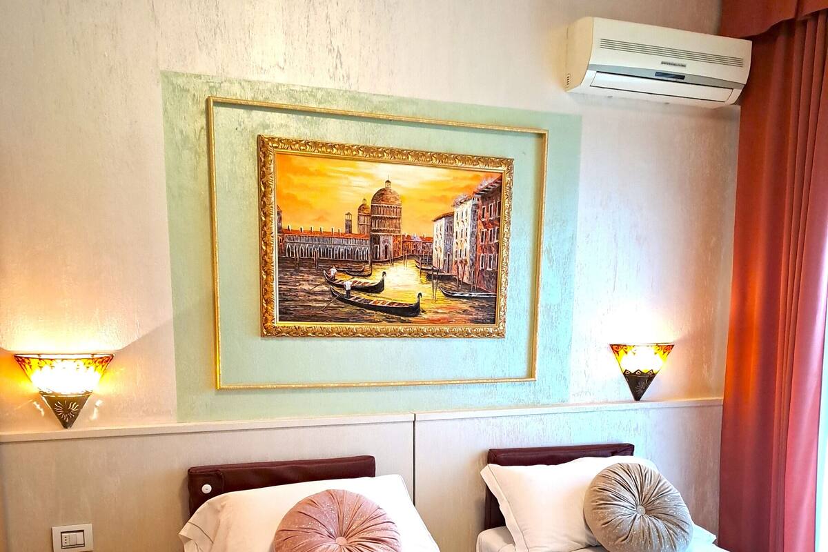 A hotel room with two beds, a painting of a cityscape, wall-mounted lights, and an air conditioning unit.