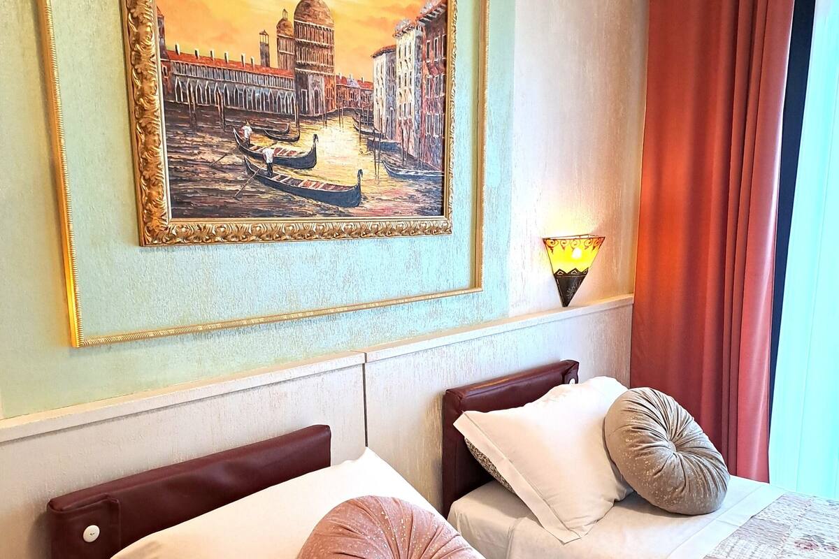 A hotel room with two beds, a painting of a cityscape, an air conditioner, and a lamp.