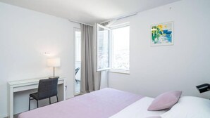 Ein Hotelzimmer mit Bett, Schreibtisch, Stuhl, Lampe, Fenster und einem Gemälde an der Wand.