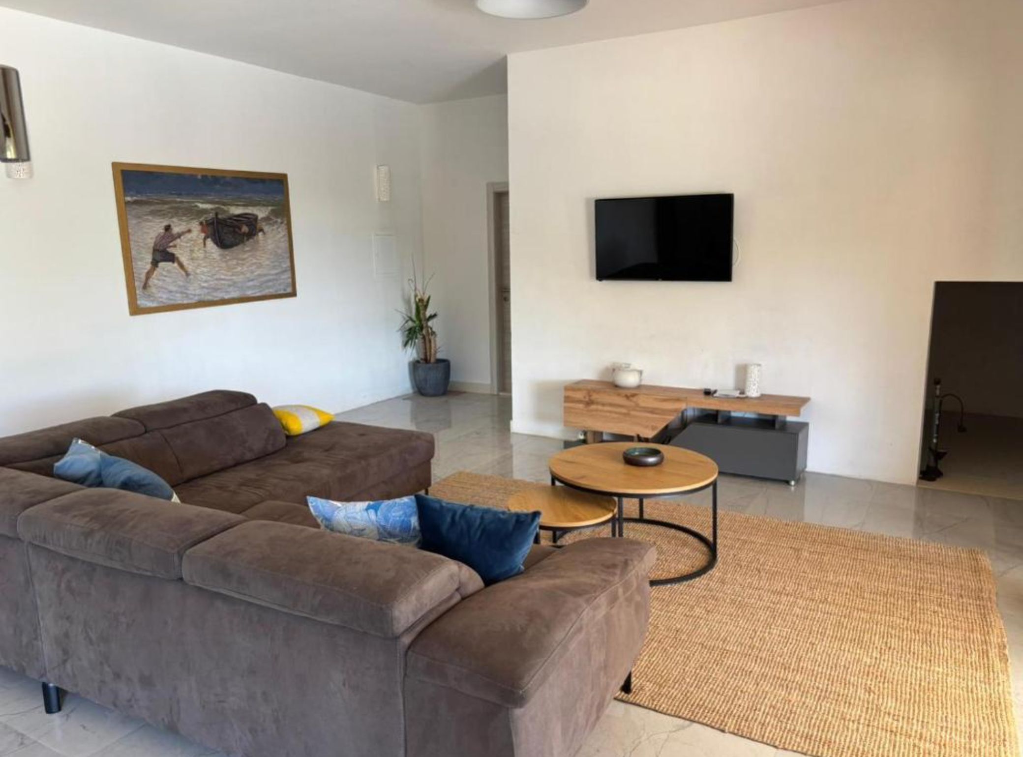 Una sala de estar moderna con un sofá marrón, una mesa de centro redonda, un televisor de pantalla plana y un cuadro con una escena de playa.