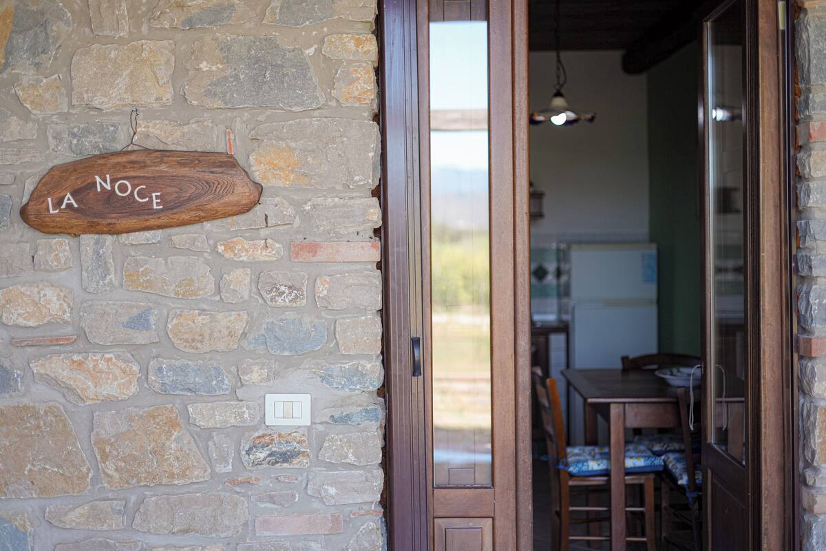 Un mur de pierre sur lequel est fixée une enseigne en bois portant l’inscription « LA NOCE », et une porte donnant sur une salle à manger avec une table et des chaises.