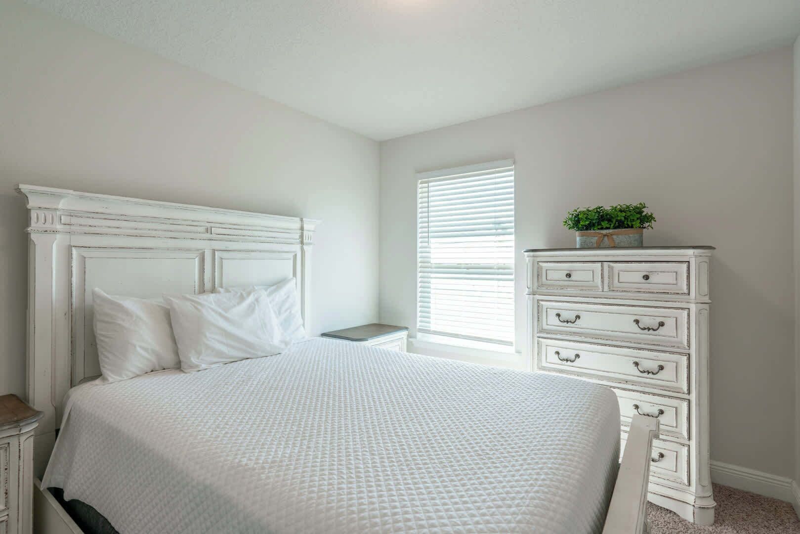 A bedroom with a white bed, a dresser, and a window with blinds.