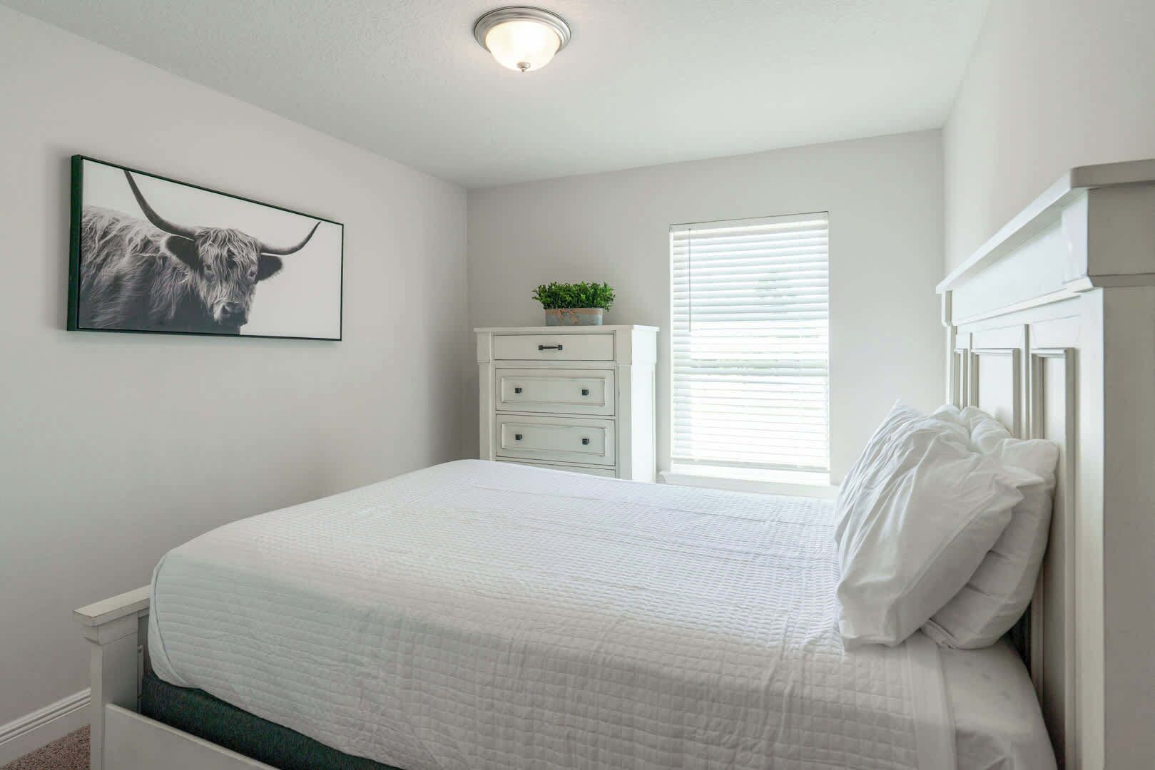 Une chambre à coucher comprenant un lit, une commode, une fenêtre avec des stores, un luminaire au plafond et un tableau encadré accroché au mur.