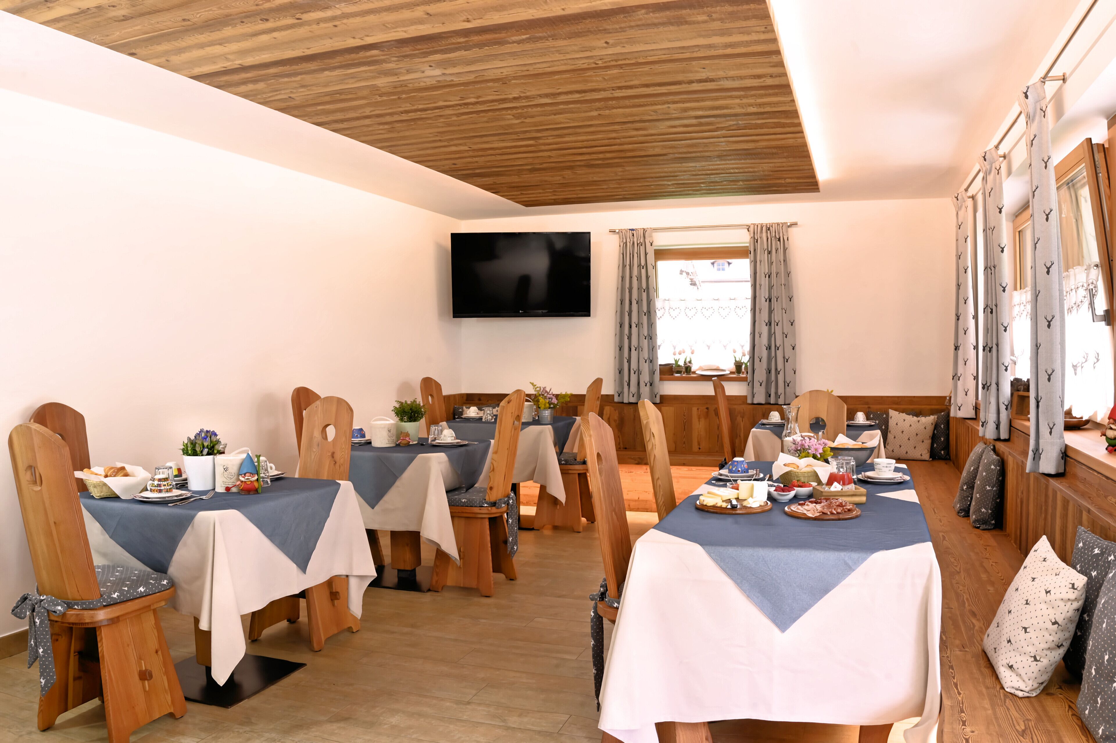 Ein Essbereich mit Holzsesseln, Tischen mit blau-weißen Tischdecken und einem an der Wand befestigten Flachbildfernseher.